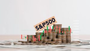 A wooden block with 'S&P 500' in black text rests diagonally on stacks of silver and gold coins. Behind and around the coins, a green and red candlestick chart is overlaid, showing market fluctuations against a light grey background with white numerical values on the right axis. Cash is visible beneath the scattered coins.