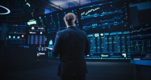 A man in a dark suit stands with his back to the viewer, observing a wall of large, glowing blue and green digital screens displaying complex financial charts, graphs, and stock market data. The room is dimly lit, highlighting the bright displays. He holds a tablet or notebook in his left hand, suggesting deep engagement with the information.