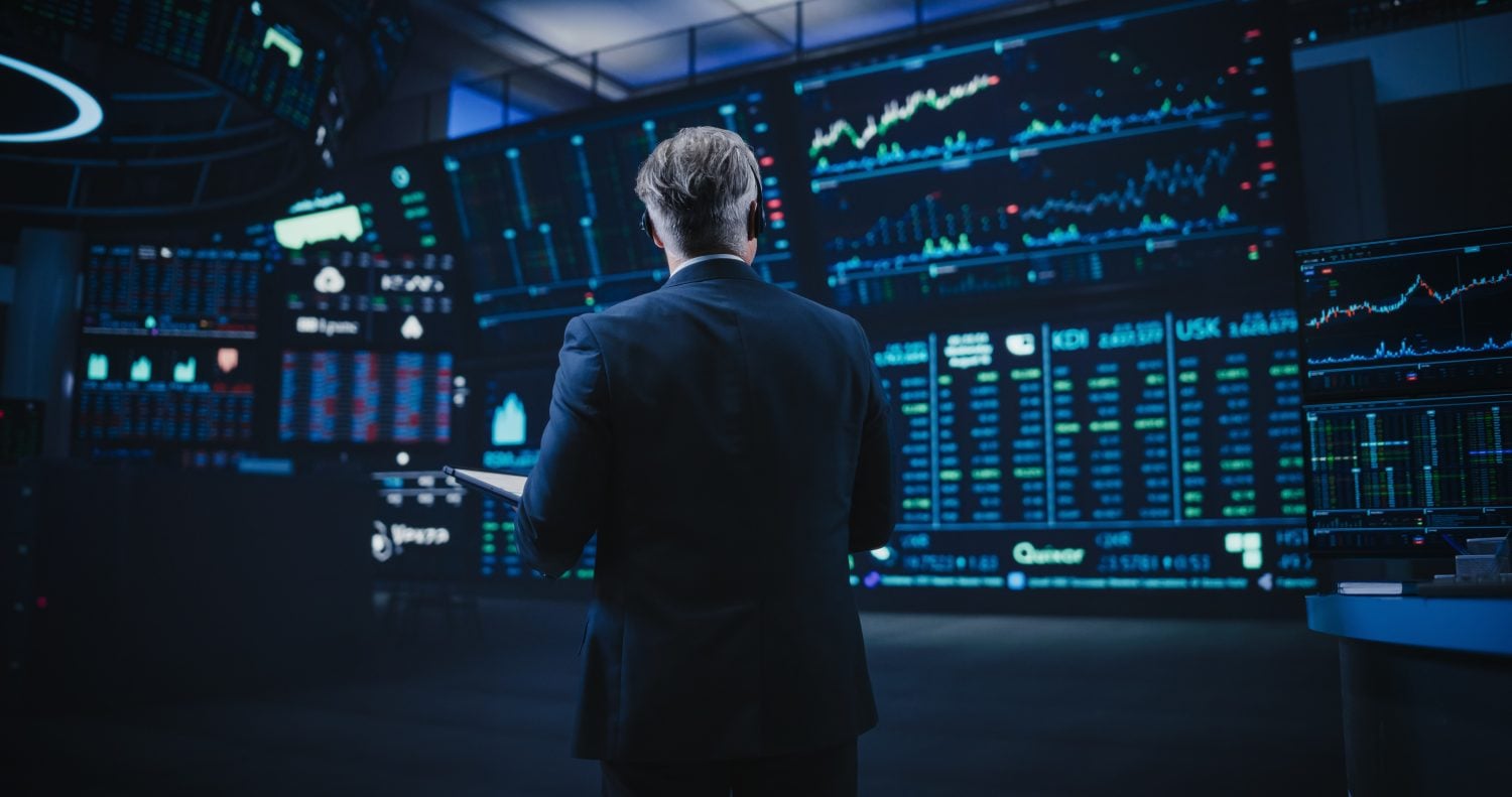 A man in a dark suit stands with his back to the viewer, observing a wall of large, glowing blue and green digital screens displaying complex financial charts, graphs, and stock market data. The room is dimly lit, highlighting the bright displays. He holds a tablet or notebook in his left hand, suggesting deep engagement with the information.