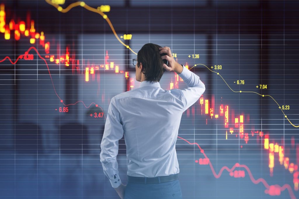A man with short dark hair and glasses, wearing a white collared shirt and dark trousers, stands with his back to the viewer. His right hand is raised, scratching the back of his head, indicating worry or contemplation. He is looking at a large, translucent holographic display that shows financial market charts with prominent red lines and red candle bars trending downwards, signifying a steep decline. A yellow line also traverses the chart, showing some fluctuations. The background is dark with a subtle grid pattern.