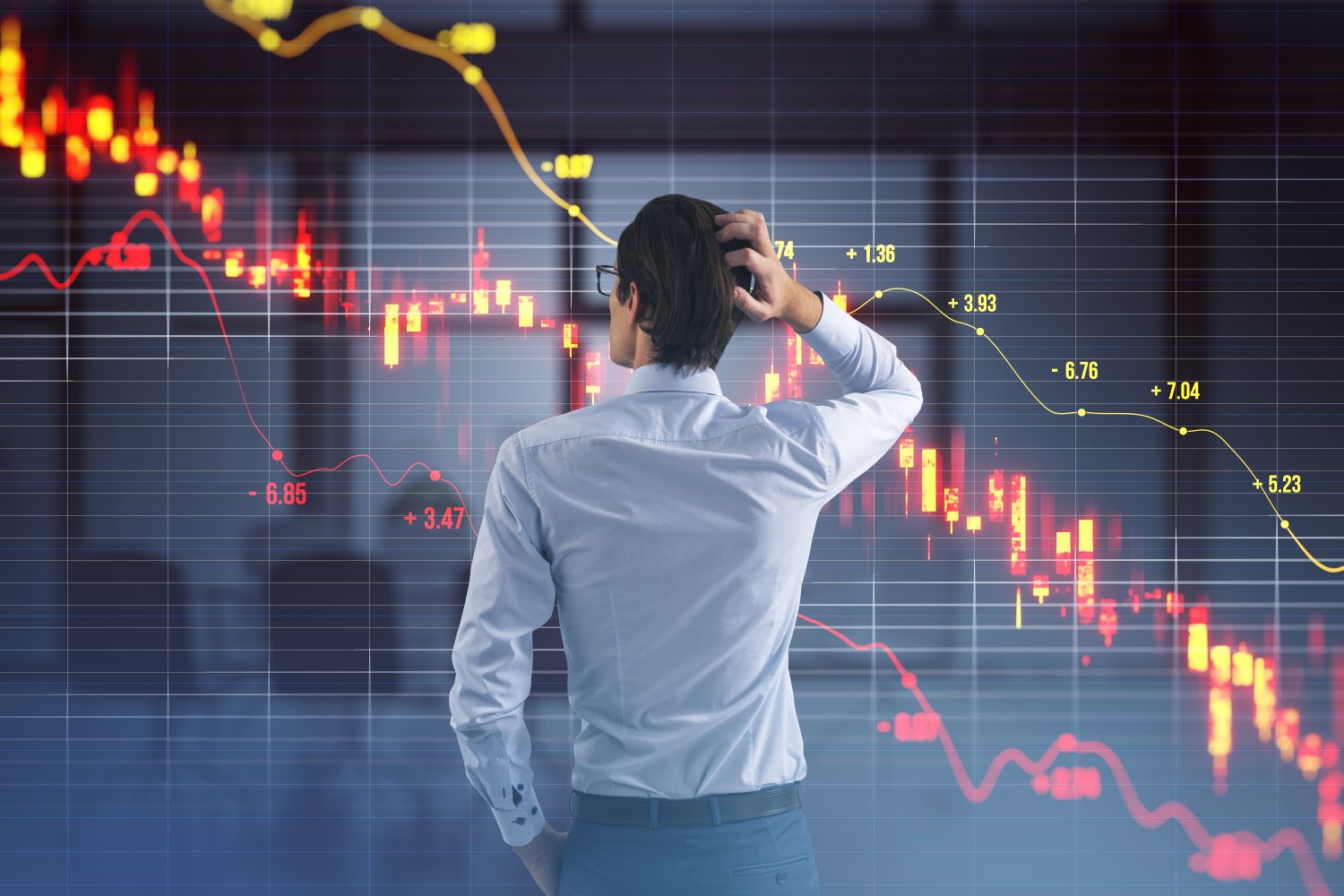 A man with short dark hair and glasses, wearing a white collared shirt and dark trousers, stands with his back to the viewer. His right hand is raised, scratching the back of his head, indicating worry or contemplation. He is looking at a large, translucent holographic display that shows financial market charts with prominent red lines and red candle bars trending downwards, signifying a steep decline. A yellow line also traverses the chart, showing some fluctuations. The background is dark with a subtle grid pattern.