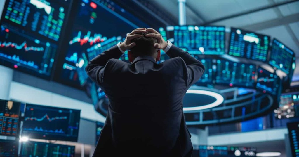 A person in a dark suit is seen from behind, with their hands clasped over their head, facing a semicircular array of glowing blue and green financial display screens. The screens show various charts, graphs, and numerical data, creating an immersive and somewhat overwhelming environment, conveying a mood of stress or deep concentration.
