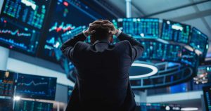 A person in a dark suit is seen from behind, with their hands clasped over their head, facing a semicircular array of glowing blue and green financial display screens. The screens show various charts, graphs, and numerical data, creating an immersive and somewhat overwhelming environment, conveying a mood of stress or deep concentration.