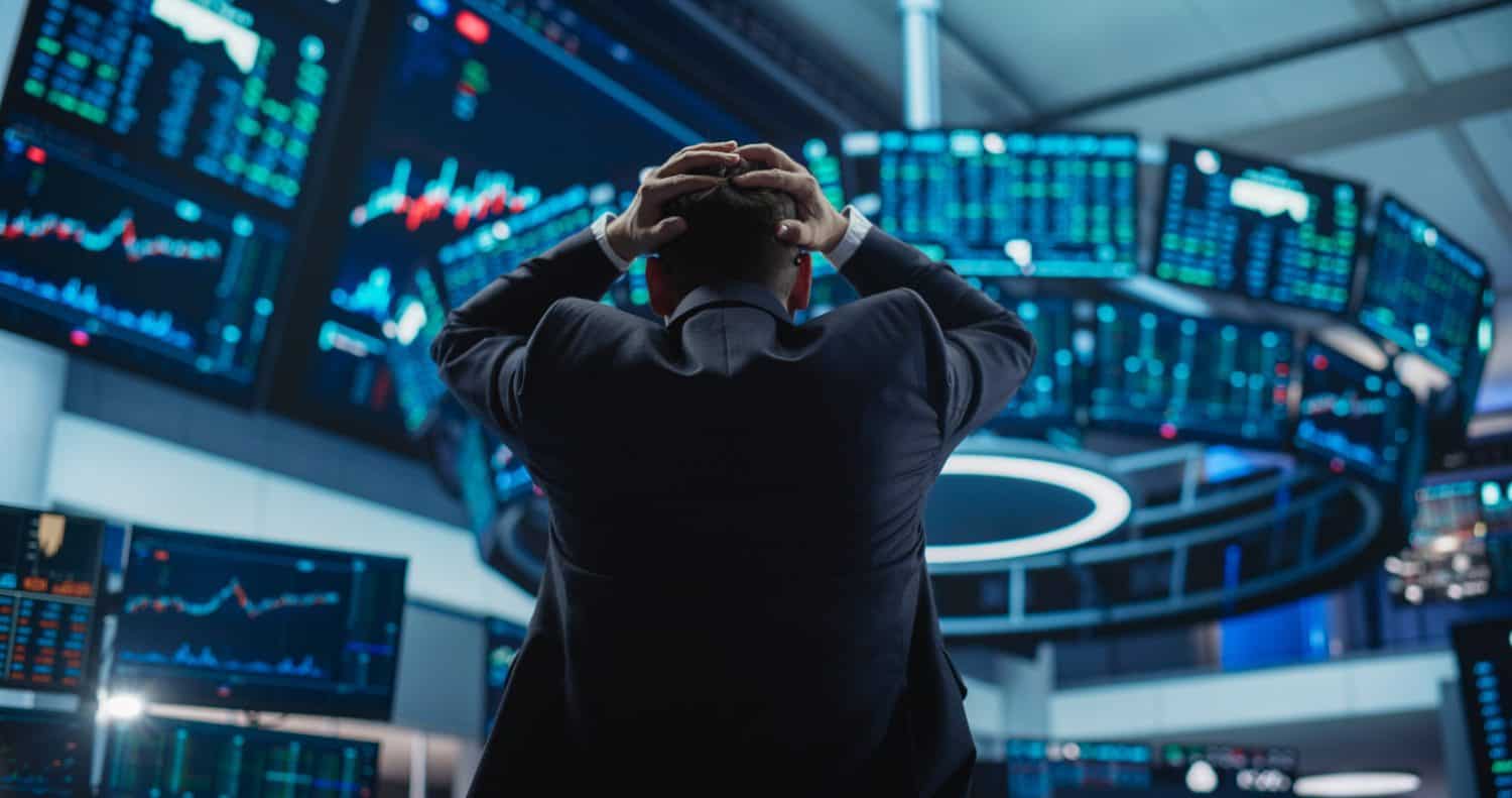 A person in a dark suit is seen from behind, with their hands clasped over their head, facing a semicircular array of glowing blue and green financial display screens. The screens show various charts, graphs, and numerical data, creating an immersive and somewhat overwhelming environment, conveying a mood of stress or deep concentration.