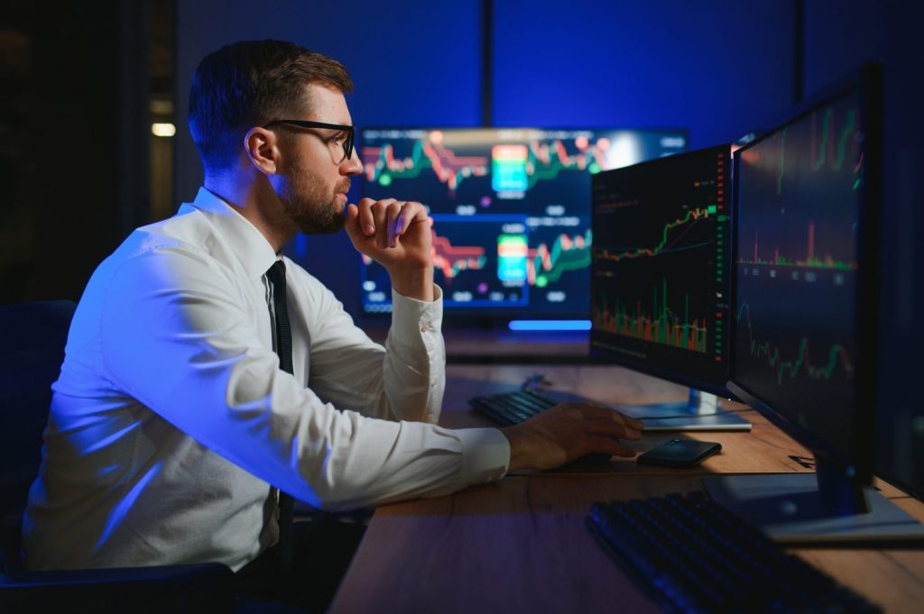A man in glasses, a white shirt, and a black tie sits at a desk in a dark room, illuminated by blue light from multiple computer monitors. He looks intently at the screens which display complex financial charts and graphs. His right hand is raised to his chin in a thoughtful pose. Keyboards and a smartphone are visible on the wooden desk.