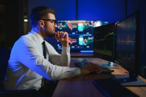 A man in glasses, a white shirt, and a black tie sits at a desk in a dark room, illuminated by blue light from multiple computer monitors. He looks intently at the screens which display complex financial charts and graphs. His right hand is raised to his chin in a thoughtful pose. Keyboards and a smartphone are visible on the wooden desk.