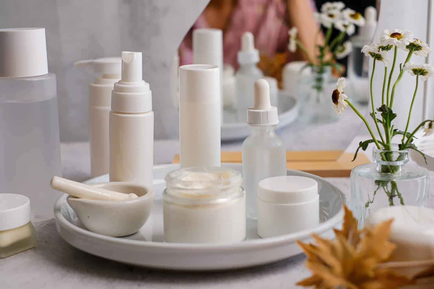 A minimalist arrangement of white and clear cosmetic and skincare products, including pump bottles, dropper bottles, jars of cream, and a mortar and pestle, displayed on a round white tray. A small glass vase with white flowers is visible in the background, along with a blurred reflection.