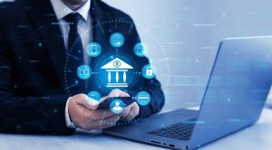 A person in a dark suit holds a smartphone, engaging with a glowing blue digital overlay representing banking and financial technology. The central icon is a bank building with a dollar sign, surrounded by interconnected icons for mobile banking, global access, financial documents, security, credit cards, user profiles, and digital receipts. A laptop is visible on the right, and the background features abstract blue digital patterns, conveying a futuristic and interconnected financial system.