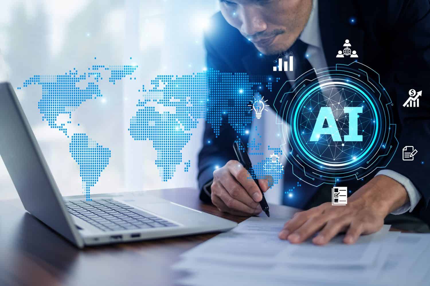 A businessman in a dark suit sits at a wooden desk, writing on a document with a black pen. An open silver laptop is to his left. Behind him, a translucent blue digital world map is overlaid with glowing white icons representing business concepts such as data analytics, finance, collaboration, innovation (lightbulb), and document management. A prominent blue circle with the white letters 'AI' is centrally featured on the right side of the image, surrounded by a complex network pattern. The scene has a professional, futuristic, and technologically advanced atmosphere.