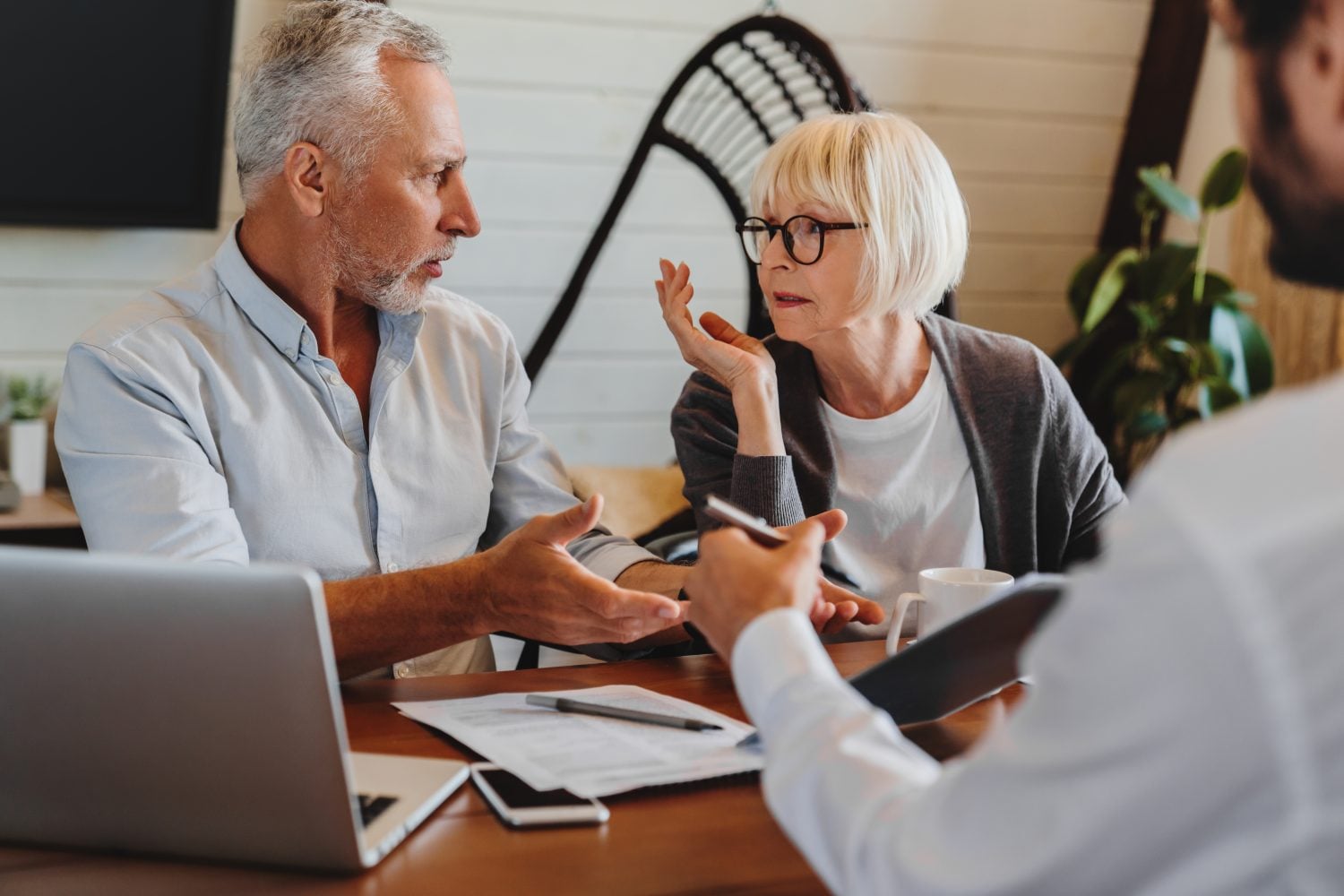A candid shot of an older couple, a man with grey hair and beard and a woman with blonde hair and glasses, in a serious discussion across a wooden table with a younger person whose back is mostly visible. A silver laptop, papers, a smartphone, and a white coffee mug are on the table. The man gestures with both hands while speaking, and the woman listens intently with a hand to her chin. The background features a light wall and a dark decorative object.