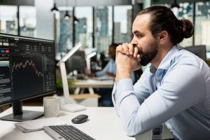 A man with a beard and hair in a bun, wearing a light blue shirt and an ID badge, sits at a white desk, looking pensively at a computer monitor displaying financial market charts with a red downward trend. His hands are clasped under his chin. On the desk are a white mug, a notepad, a black computer mouse, a silver keyboard, and a white modern desk lamp. The background shows a blurred office with other workers and city buildings through large windows.