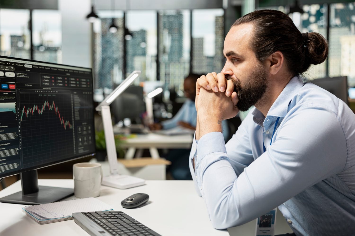 A man with a beard and hair in a bun, wearing a light blue shirt and an ID badge, sits at a white desk, looking pensively at a computer monitor displaying financial market charts with a red downward trend. His hands are clasped under his chin. On the desk are a white mug, a notepad, a black computer mouse, a silver keyboard, and a white modern desk lamp. The background shows a blurred office with other workers and city buildings through large windows.