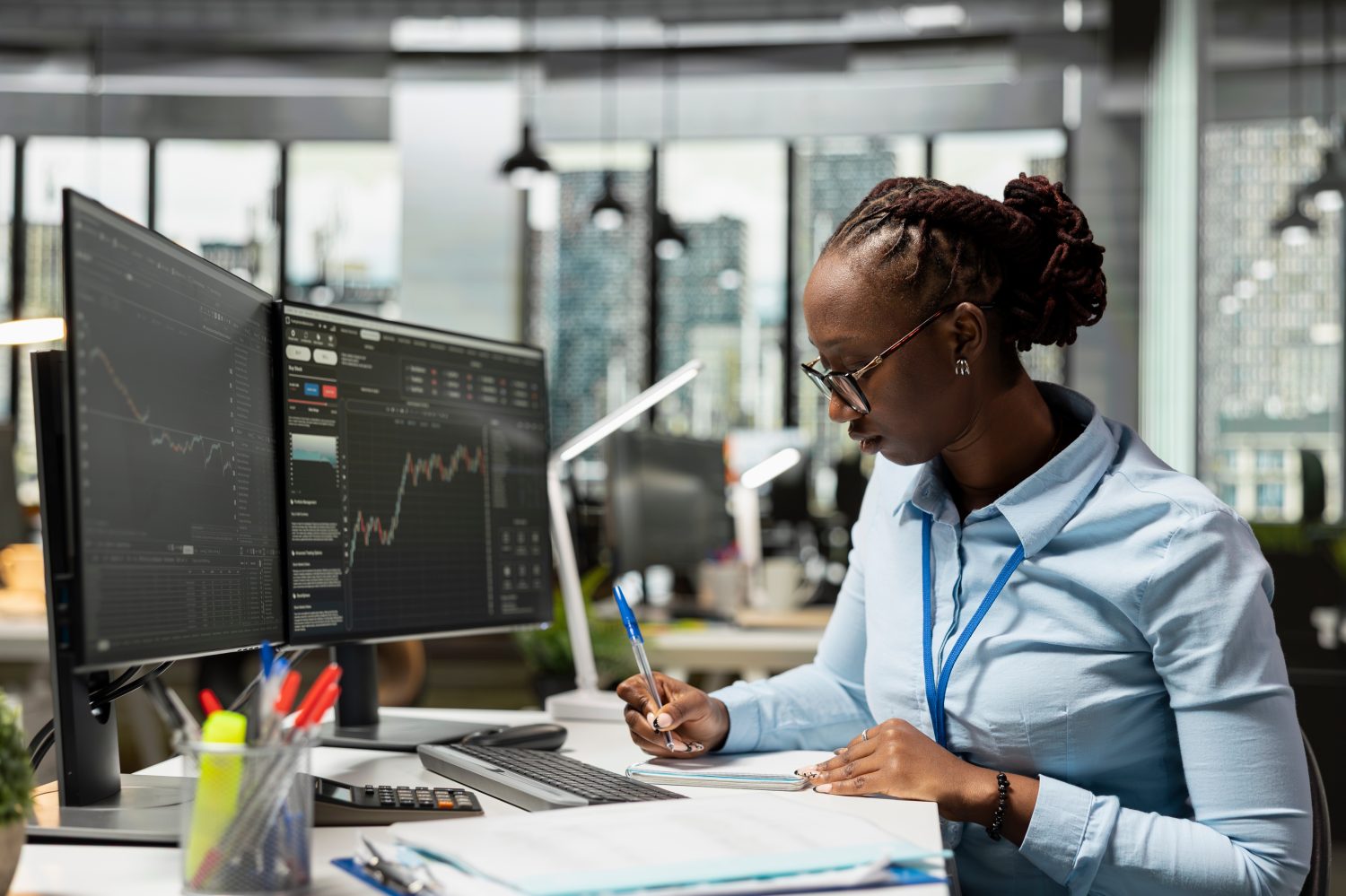 A Black woman with dreadlocks pulled back and wearing glasses sits at a white desk in a modern office, writing in a spiral-bound notebook with a blue pen. To her left, two large computer monitors display dark interfaces with colorful line graphs, stock data, and financial charts. She wears a light blue collared shirt and a blue lanyard. On the desk, there is a clear cup holding red and blue pens, a yellow highlighter, a black calculator, a keyboard, and stacks of papers.