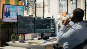 Viewed from behind, an African American man in a light blue striped shirt sits at a white desk, holding a beige corded telephone to his ear and gesturing with his left hand. In front of him are two dark-themed computer monitors displaying detailed stock charts and financial metrics. On the desk, a small potted green plant and a pen holder filled with colorful pens are visible. In the background, a large television screen shows two news presenters, and through large office windows, blurred city buildings can be seen.