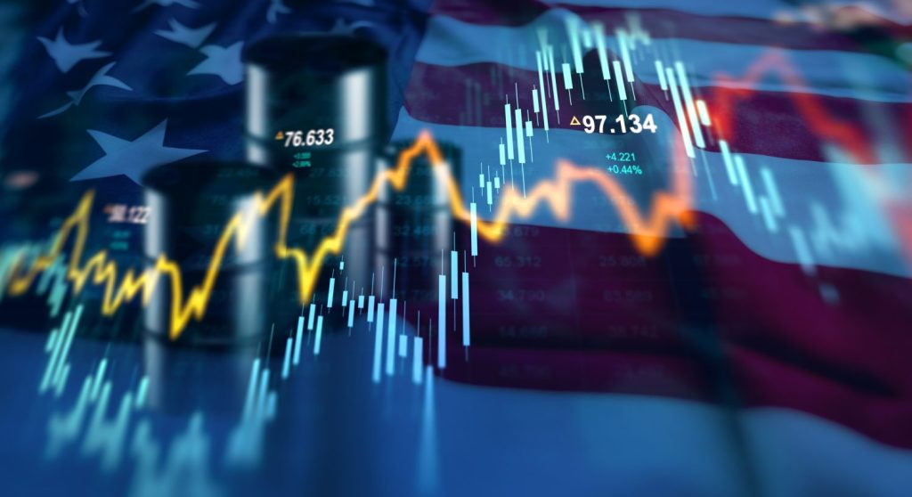 A vibrant digital overlay of financial charts, including blue candlestick bars and a bright yellow line graph, on a softly blurred American flag background. Two dark oil barrels are faintly visible behind the charts. Several numbers, such as '97.134', '76.633', '+4.221', and '+0.44%', are displayed on the graphs, suggesting active market data.