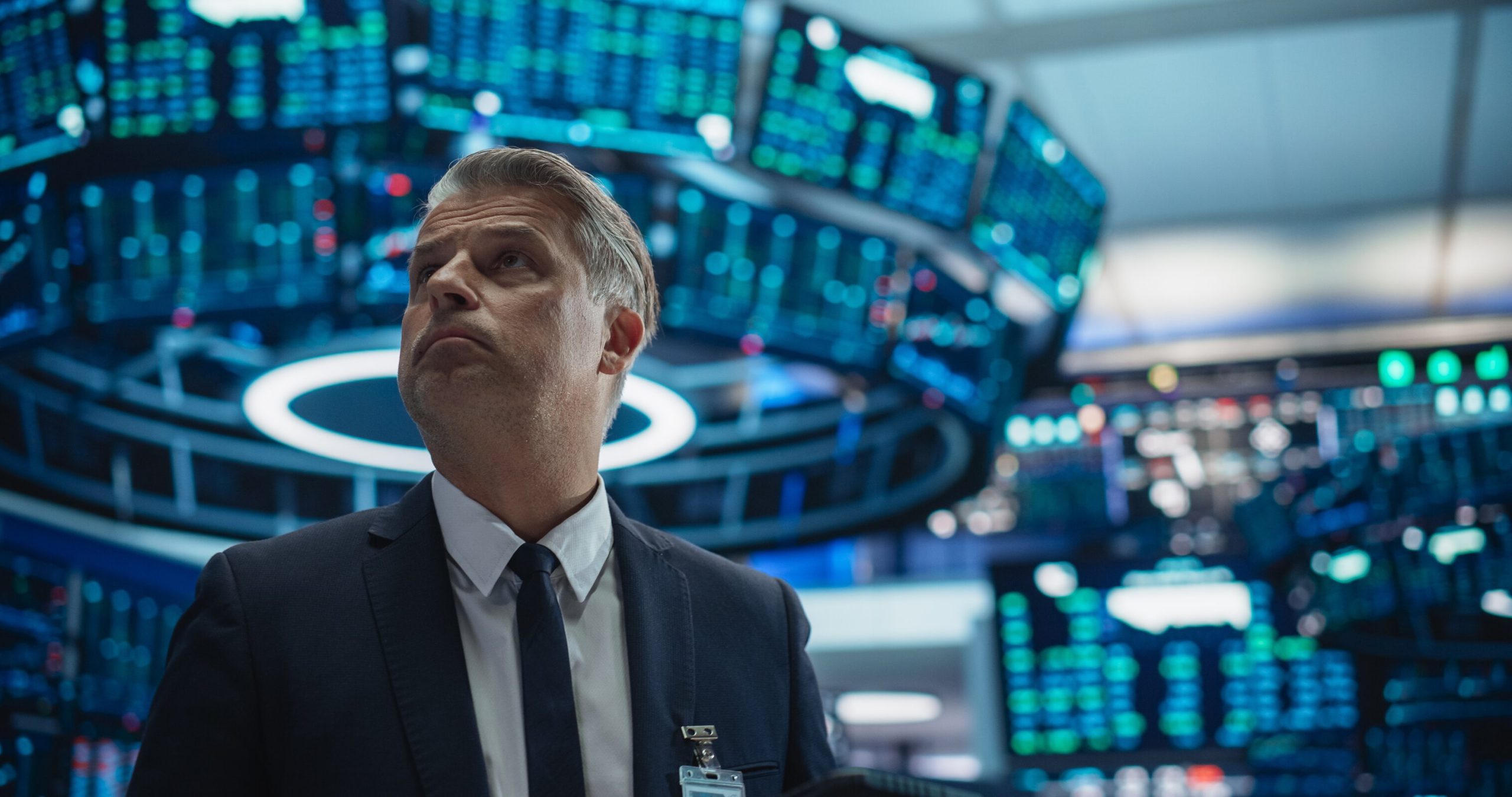 A middle-aged man with graying hair, wearing a dark suit and tie, looks upwards with a serious expression. He stands in the foreground, slightly to the left. The background is blurred, featuring numerous glowing blue and green digital screens displaying financial data, suggesting a high-tech control room or trading floor.